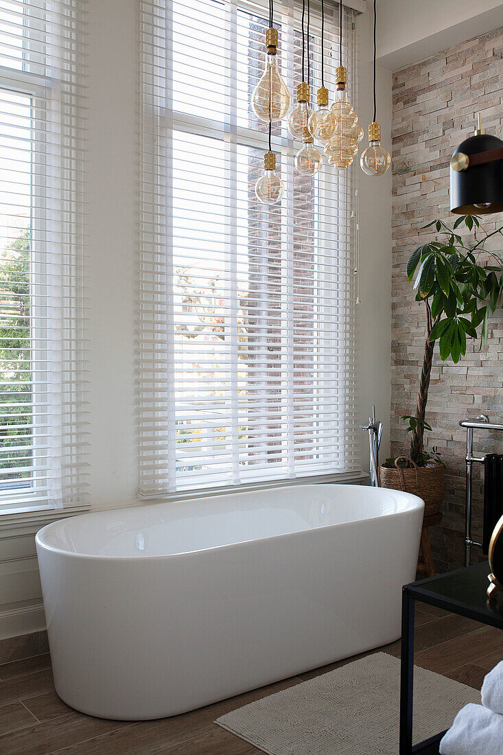 Freestanding bathtub in front of large windows with blinds and pendant lights