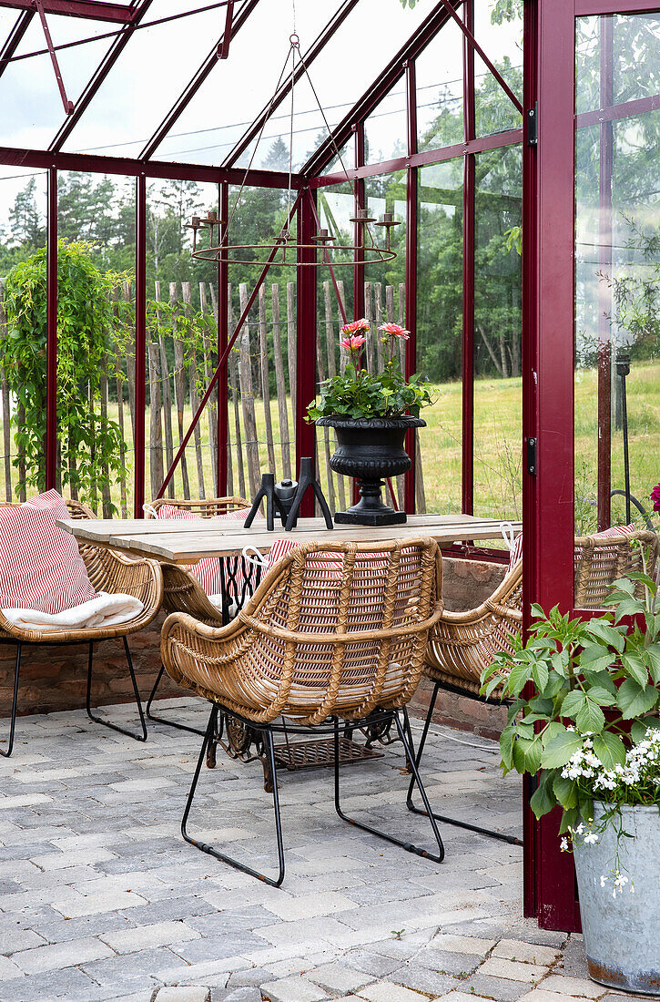 Seating area in the red glass house with rattan armchairs and wooden table on a paved floor