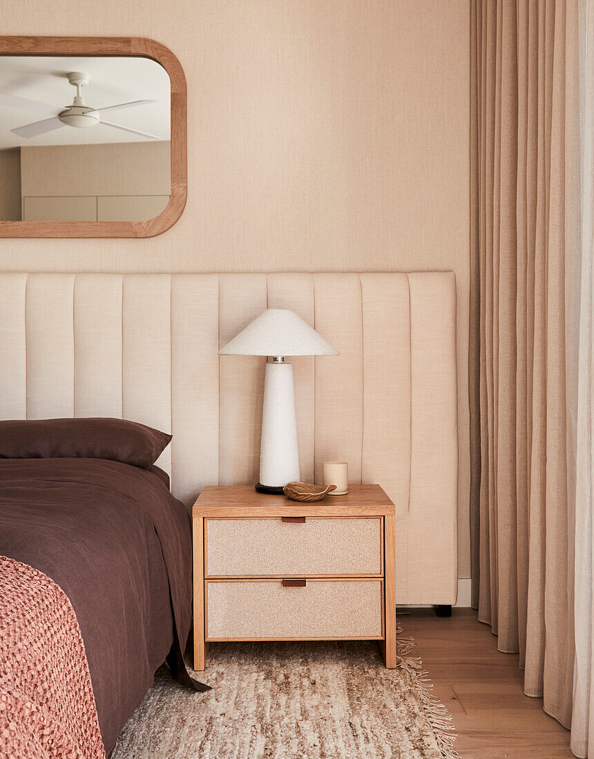 Wooden bedside table with lamp next to upholstered bed headboard in neutral bedroom