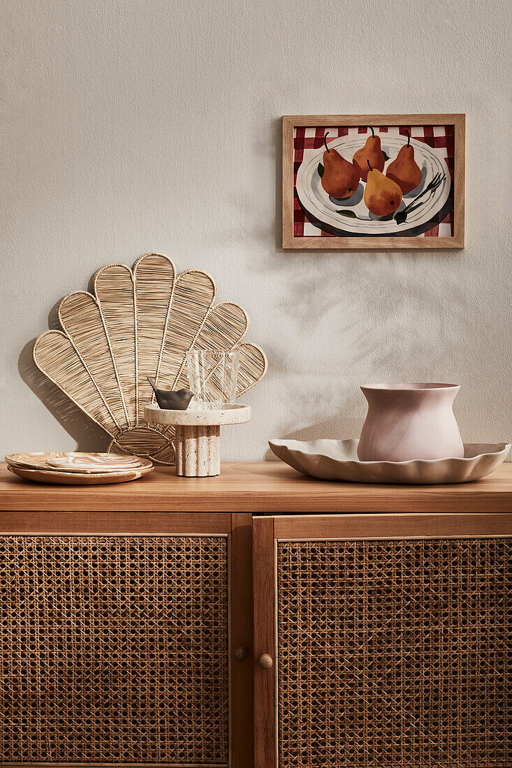 Wooden chest of drawers with rattan weave and ceramic decoration in front of framed pear picture