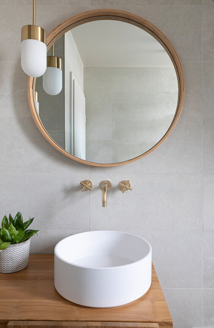 Round mirror with wooden frame above modern washbasin with golden fittings