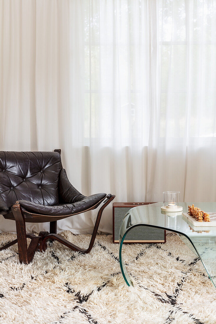 Leather chair and glass table on deep-pile carpet in front of light-coloured curtains