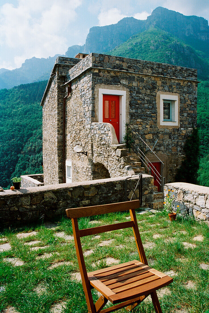 Steinhaus mit roter Tür und Gartenstuhl vor Bergkulisse