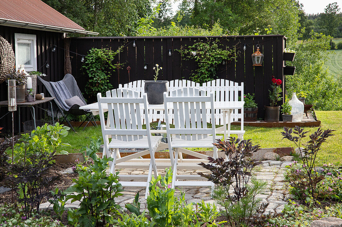 Weiße Gartenmöbel auf gepflasterter Terrasse vor schwarzem Holzzaun und grüner Landschaft