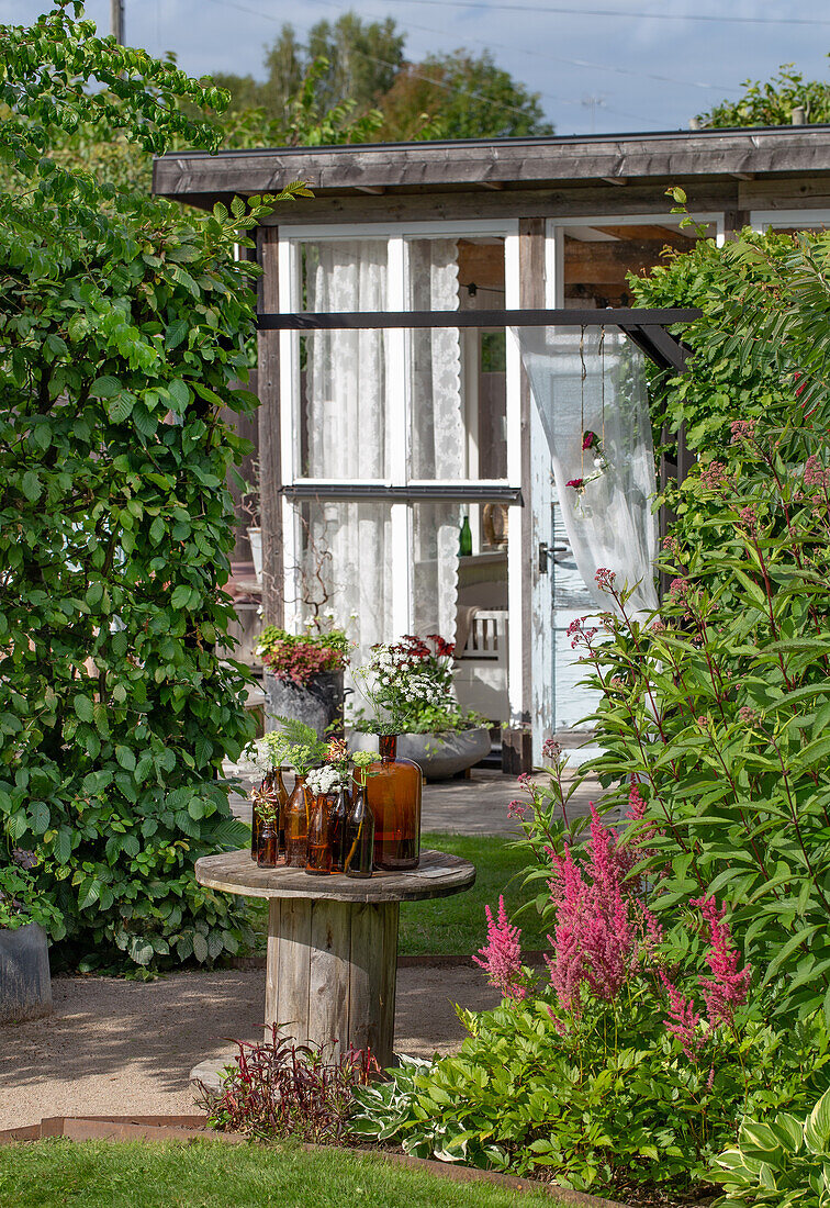 Gartenidylle mit Glasflaschen auf rustikalem Holztisch vor Gartenhaus