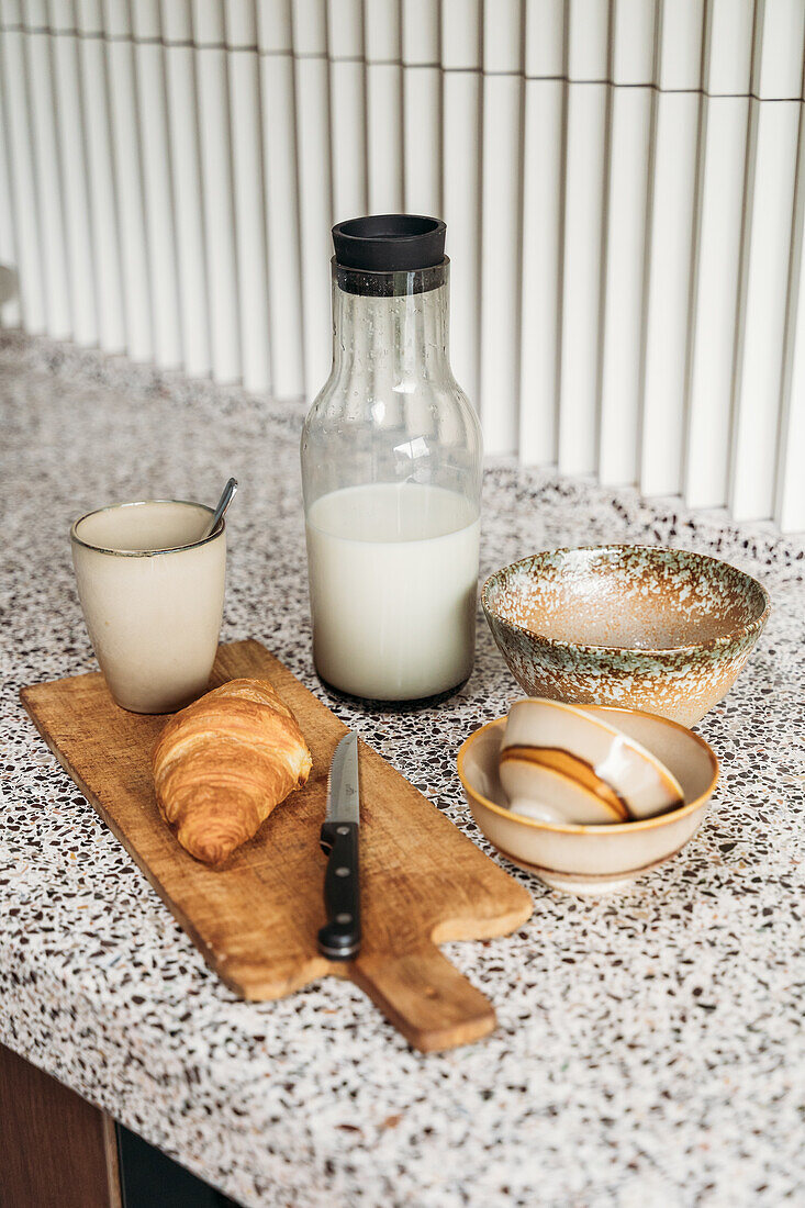 Frühstücksarrangement aus Croissant, Milchflasche und Keramikgeschirr auf der Küchenarbeitsplatte