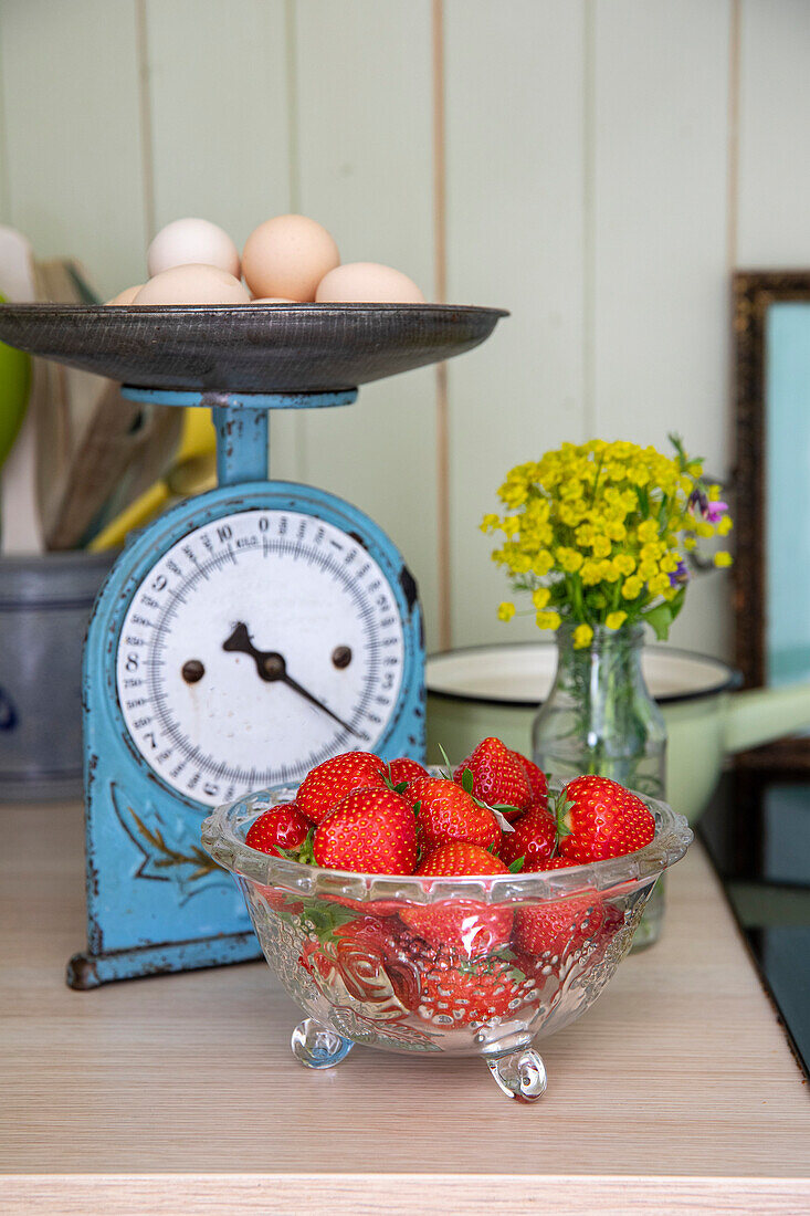 Glass bowl with strawberries next to blue scale with eggs and vase with yellow flowers