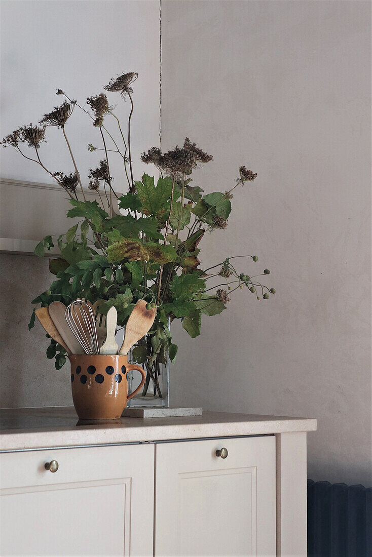 Ceramic pot with kitchen utensils and dried flowers on cream-colored cupboard