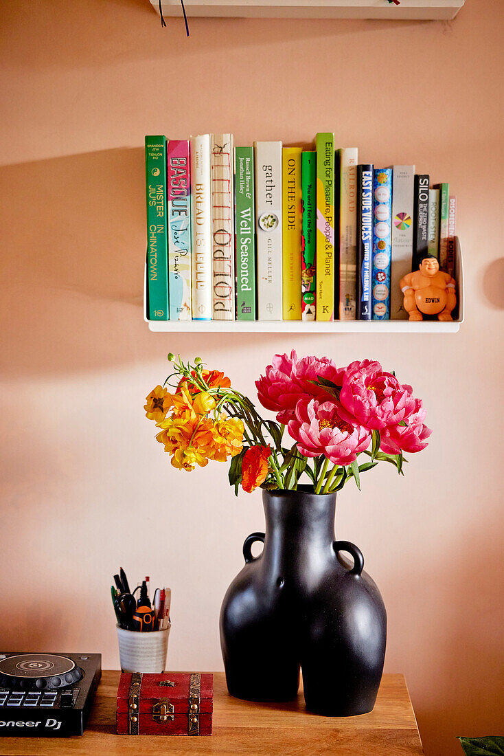 Black vase with flowers on a wooden table in front of a bookshelf, bouquet of peonies (Paeonia)