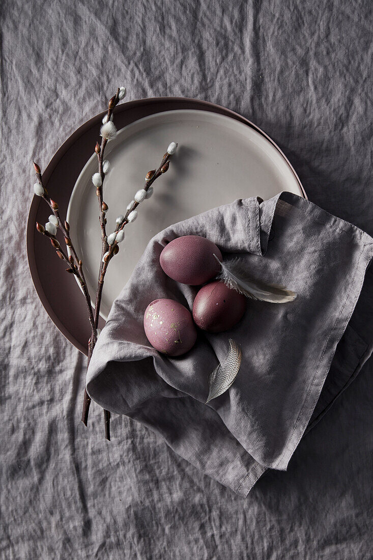 Purple Easter eggs with feathers on a grey linen cloth, pussy willows on a plate