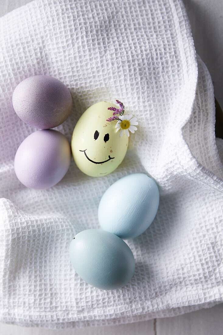 Pastel-colored Easter eggs and yellow Easter egg with painted smiling face and flowers on white cloth