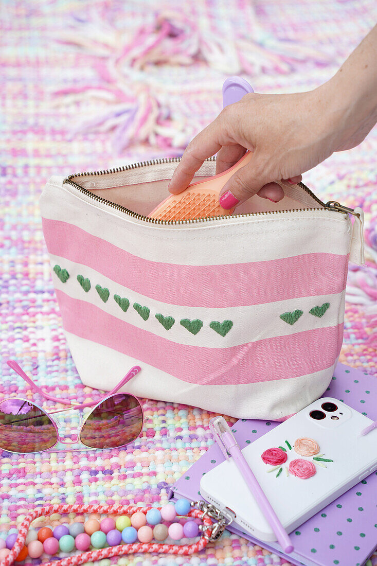 Cosmetic bag with pink stripes and green hearts, sunglasses and mobile phone on a colourful blanket