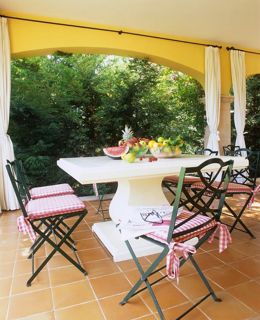 Verschiedene Früchte auf Steintisch mit geschwungenem Säulenfuss und Stühle im Landhausstil auf einer Terrasse