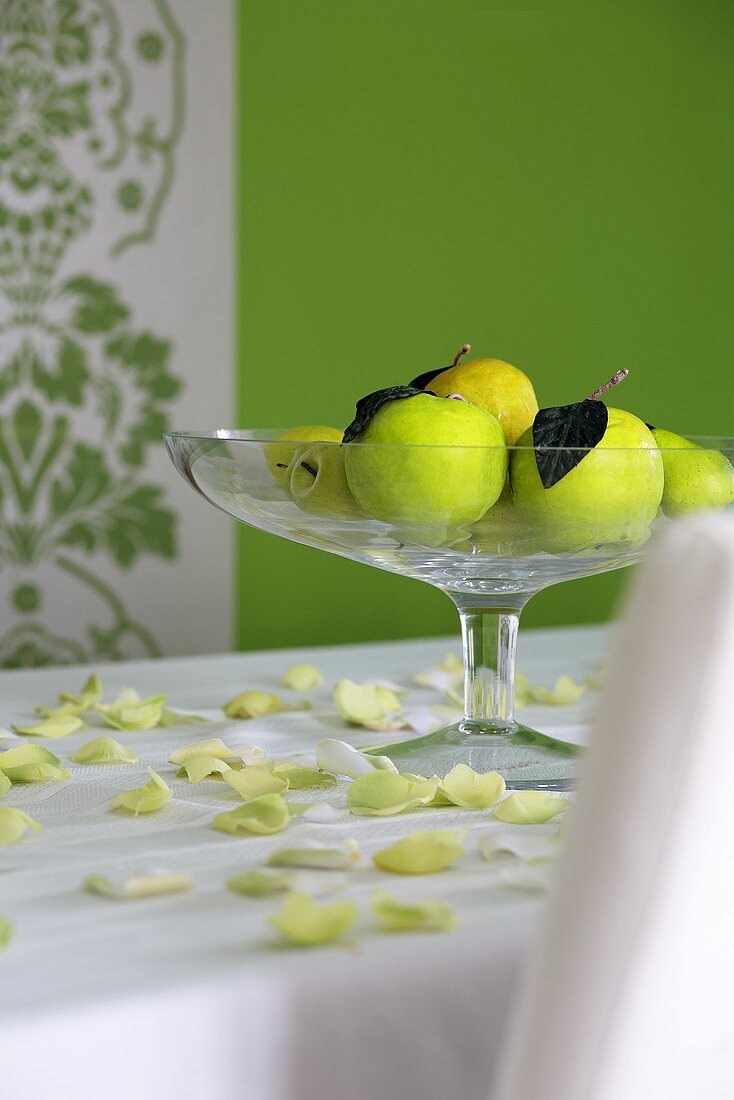 Green apples in glass fruit bowl on white table cloth scattered with yellowy-green petals