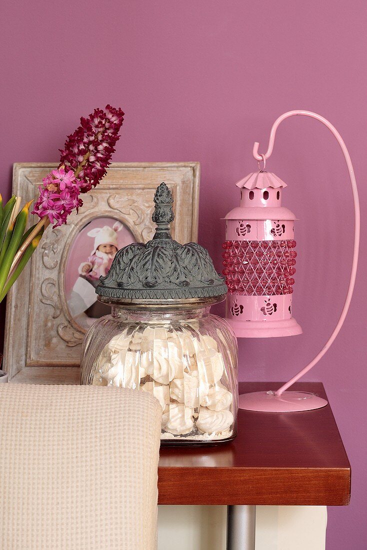 Pink lantern, glass jar of meringues and framed baby photo against lilac wall