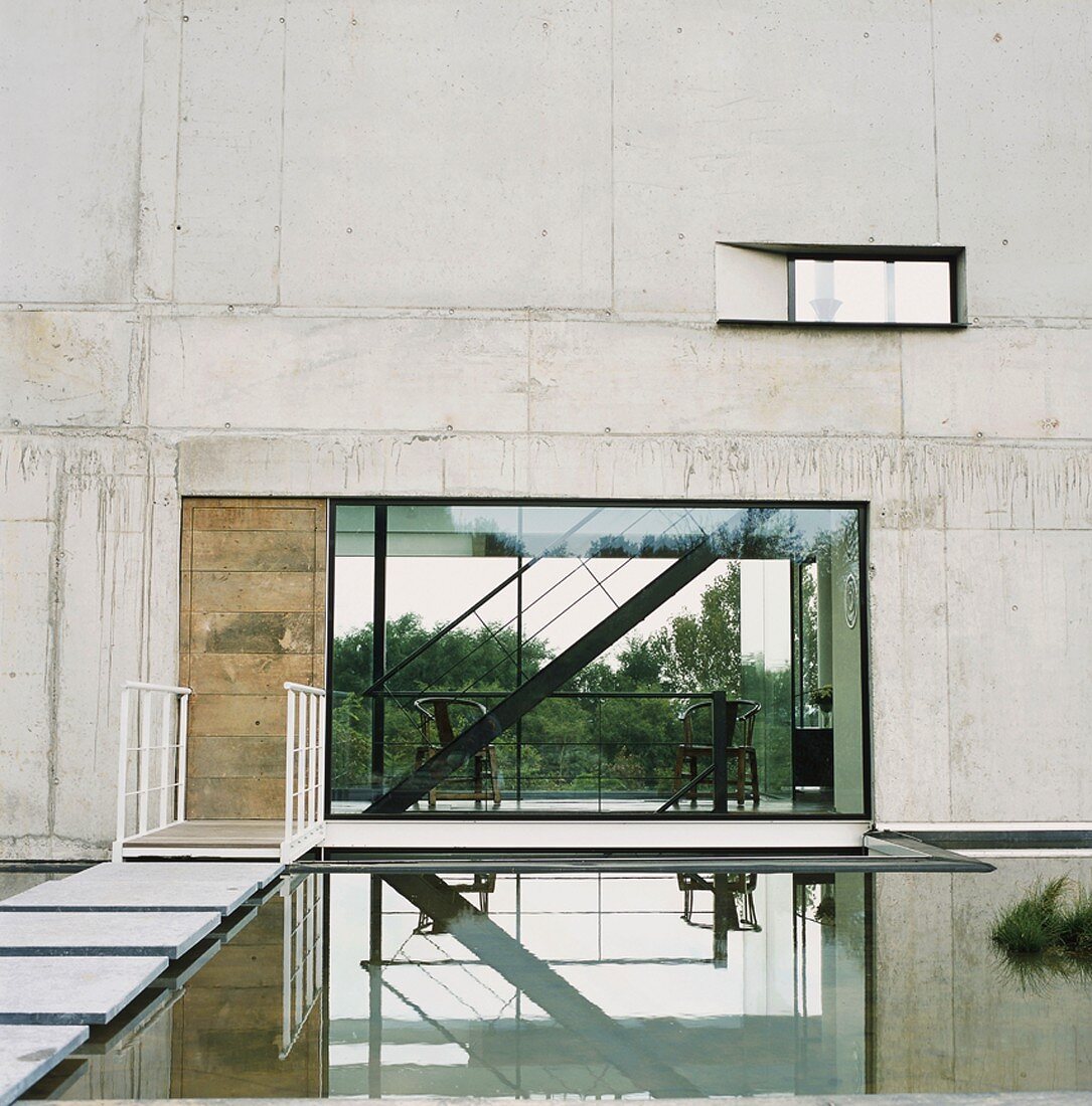 Zeitgenössische Betonarchitektur - Wasserbecken mit Steg und Einblick in offenen Wohnraum mit freistehender Treppe