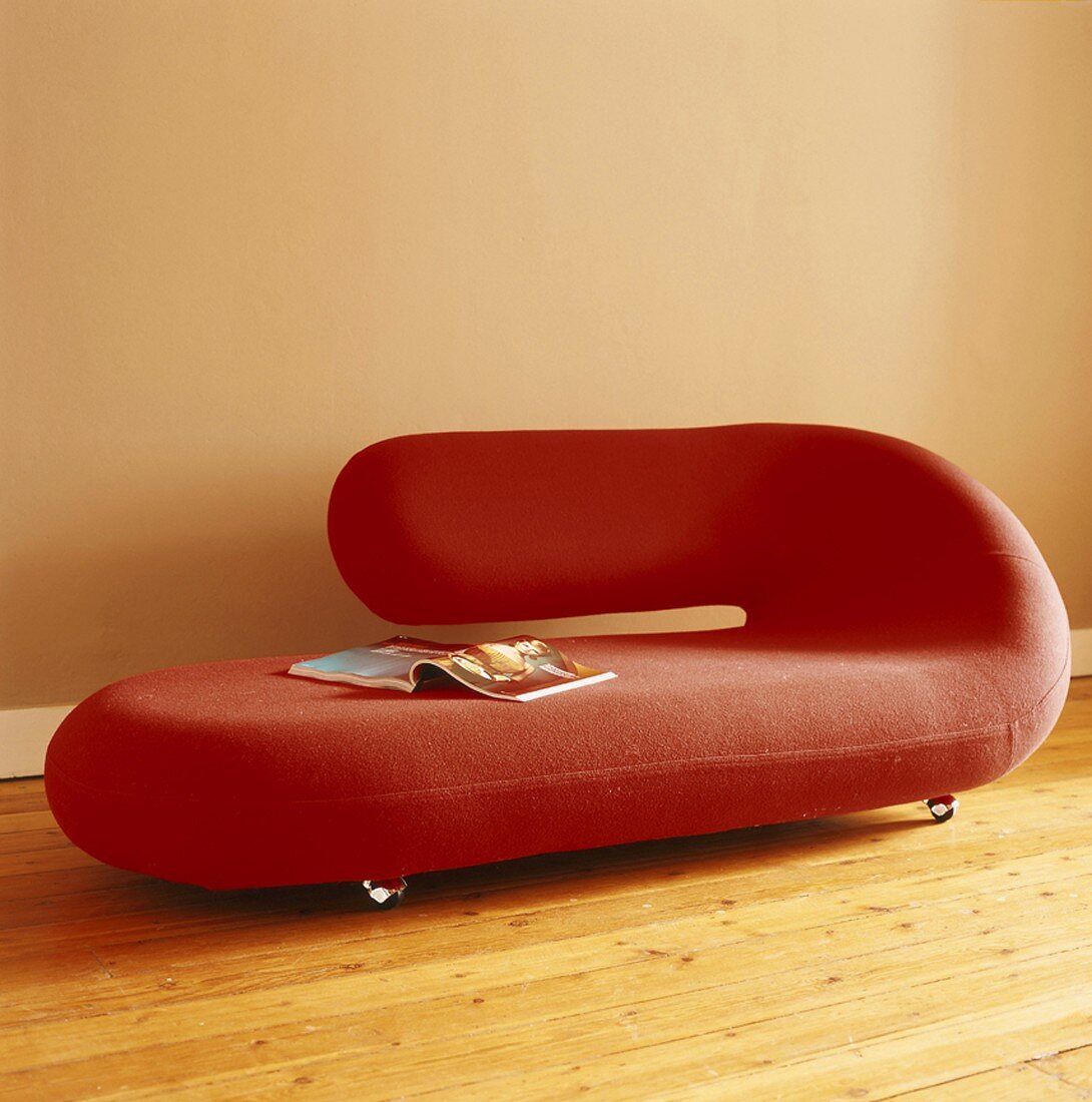 Rotes, gerundetes Designer-Sofa auf Rollen vor beigefarbener Wand