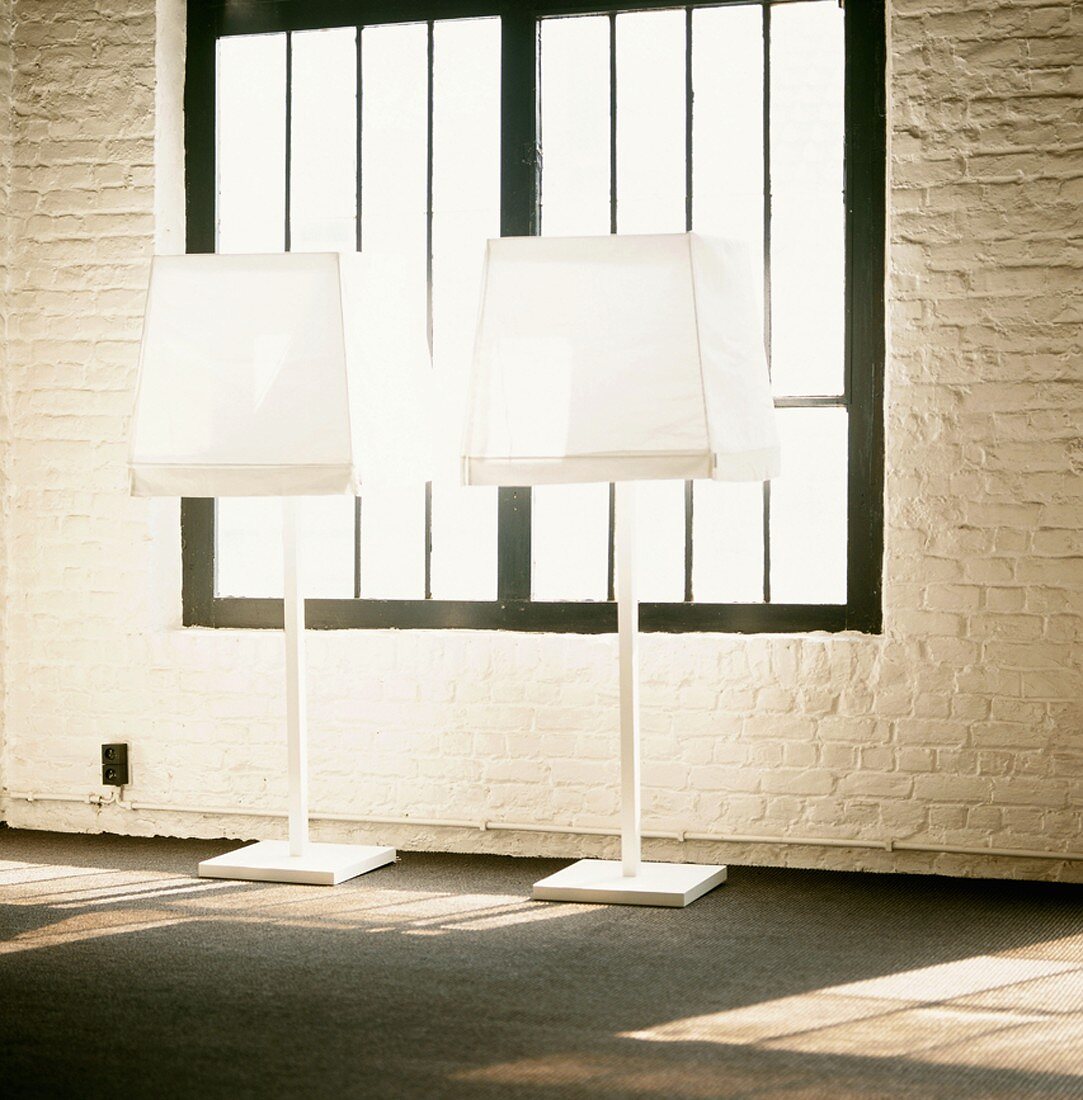Schwarzgerahmtes, altes Fabrikfenster und geweisselte Ziegelwand hinter weissen Stehlampen