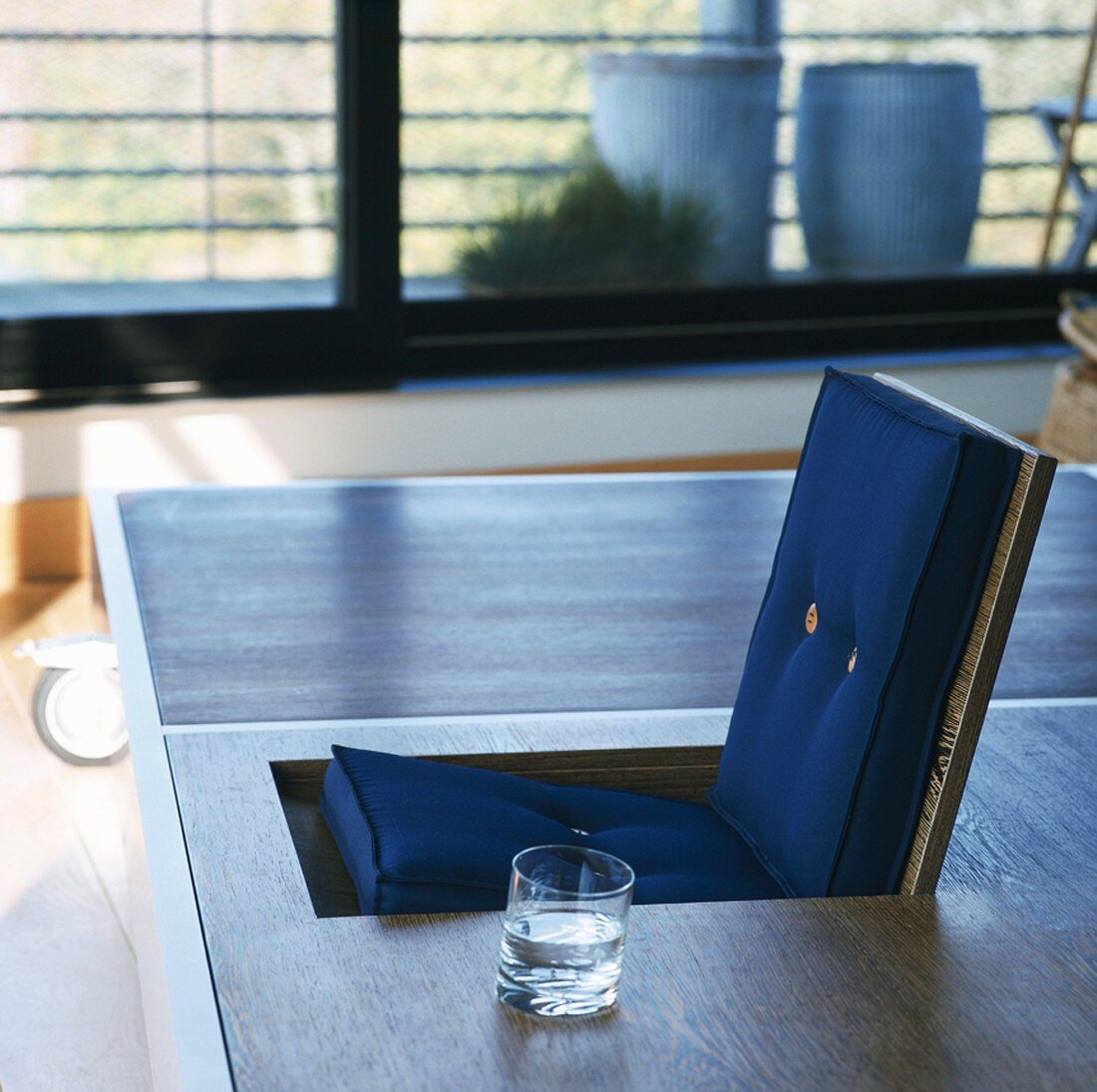 Glass of water on designer table with integrated folding chair; large, modern sliding glass doors in background