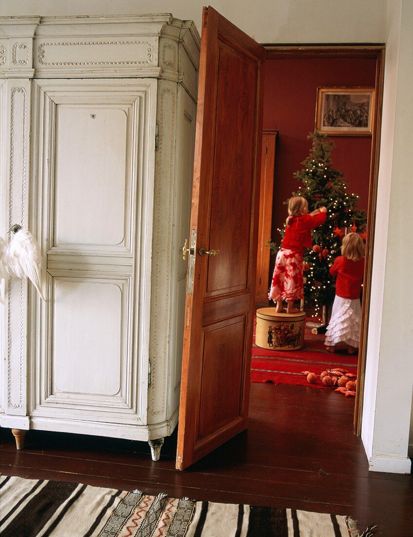 Alter Kleiderschrank im weissen Vintage-Look und Blick durch Tür auf Kinder beim Schmücken des Weihnachtsbaums