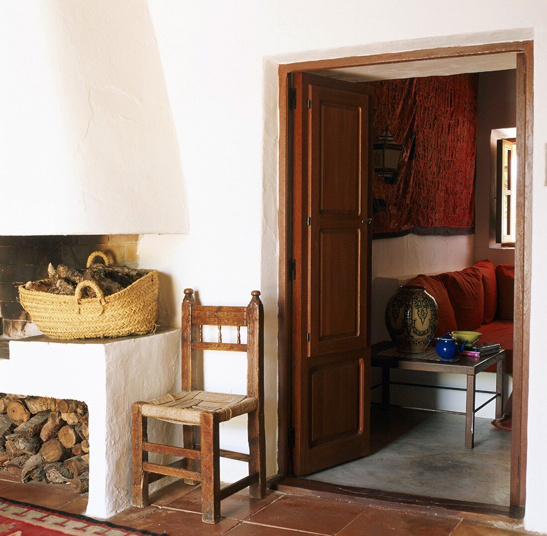Weiß verputzter Kamin mit Holzvorräten und Blick in gemütlichen Wohnraum mit Kissen auf gemauertem Sofa