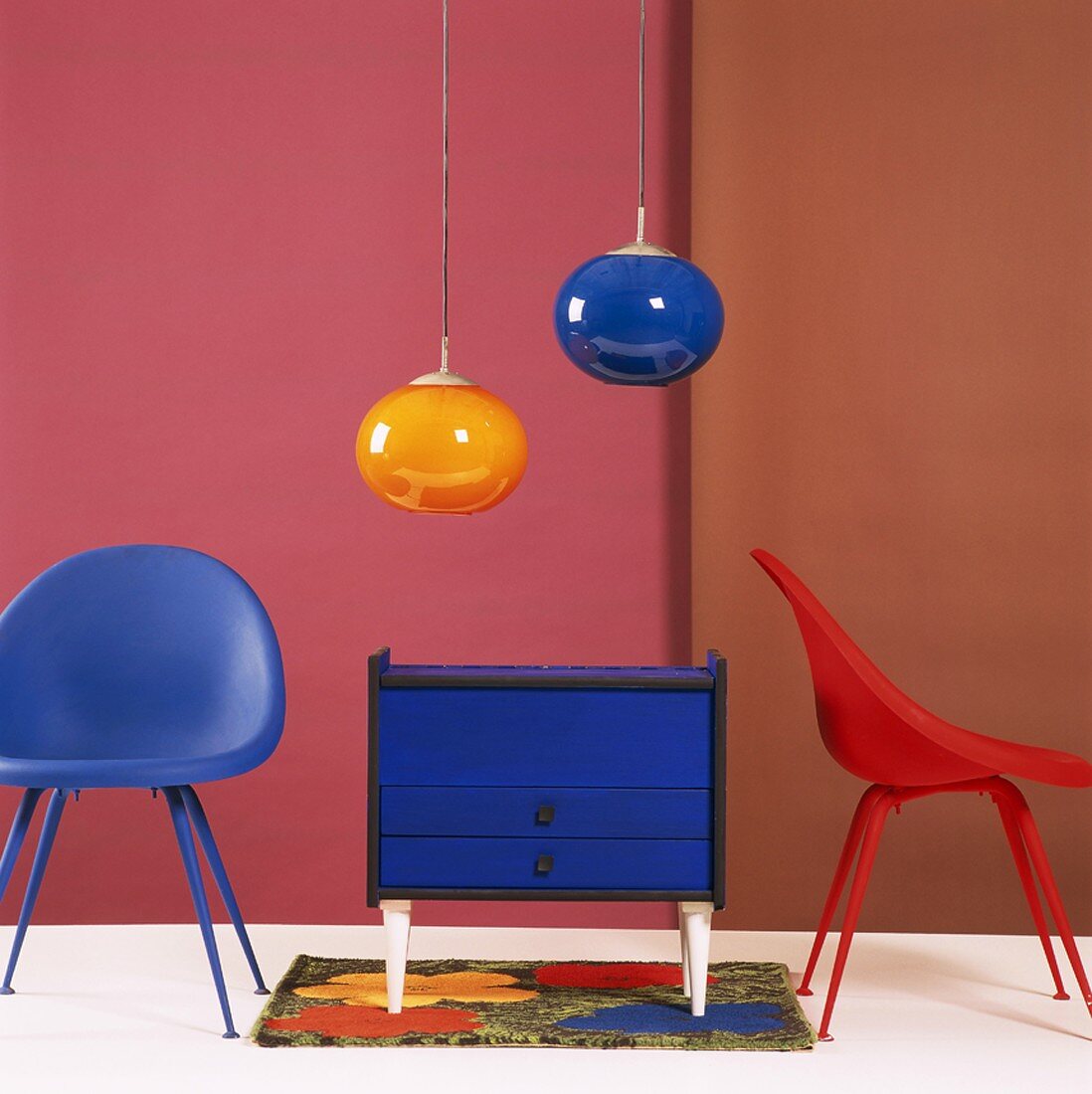 Kleine blaue Kommode zwischen Kunststoff-Schalenstühlen und zwei Retro-Hängelampen aus farbigem Glas