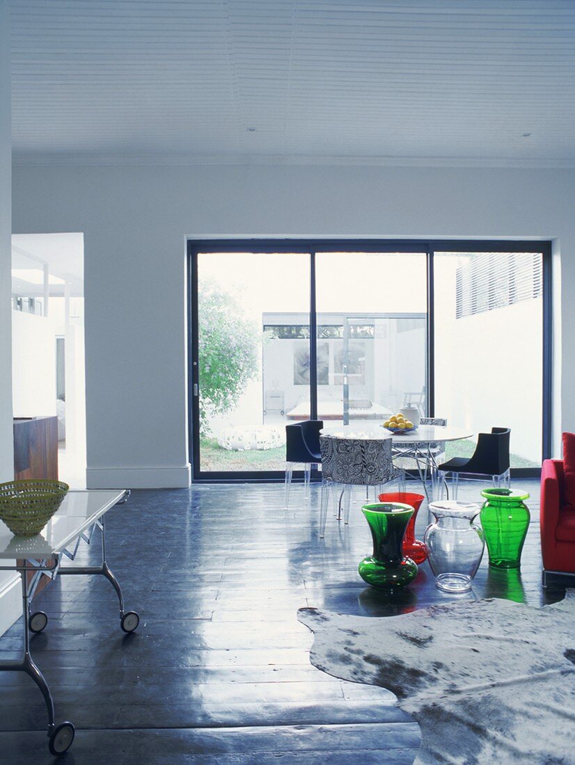 Sparsam möblierter offener Wohn/Essraum mit Esstisch vor der Fensterfront und farbigen Glasvasen auf dem Boden
