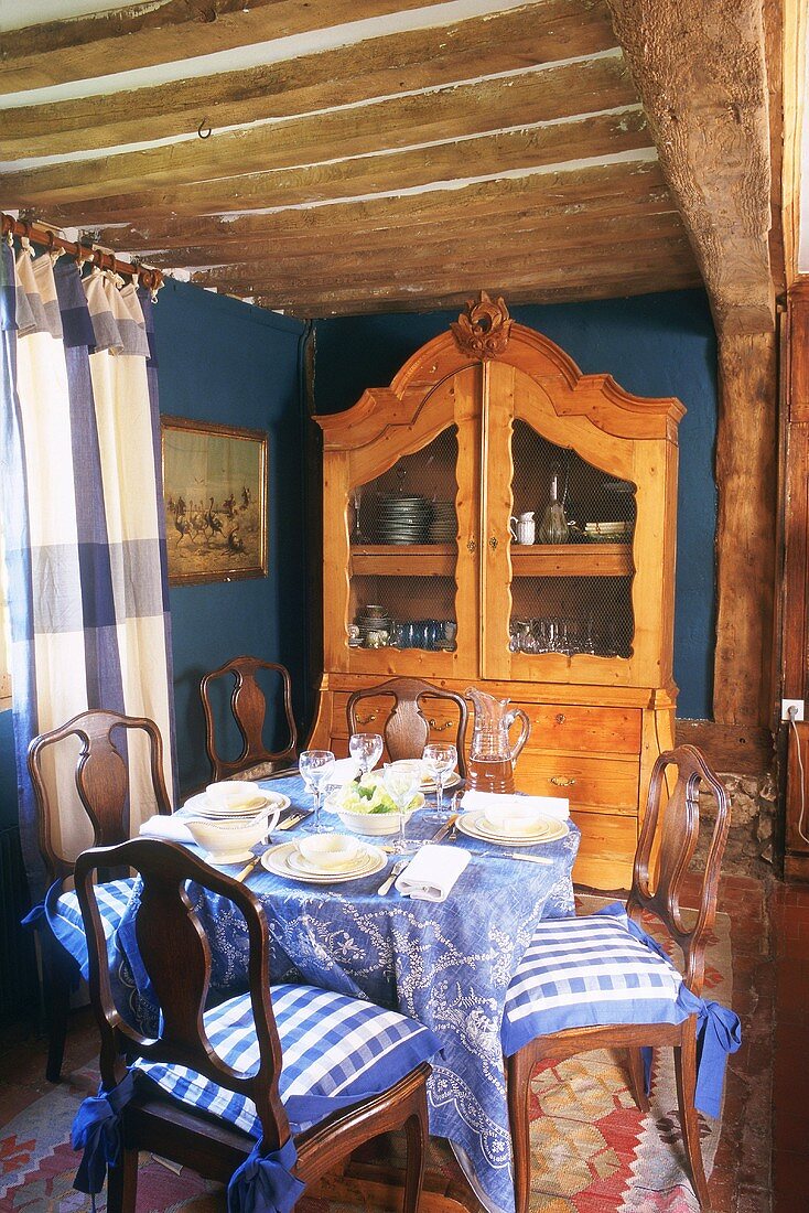 Blue dining room with set table, blue and white checked curtains and wood-beamed ceiling