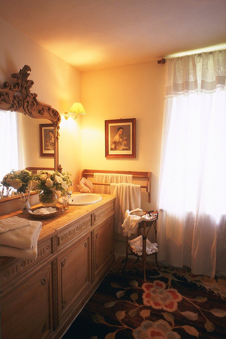 Traditional bathroom with wooden cabinet as washstand; large, framed mirror; floral carpet and translucent curtain with lace edging