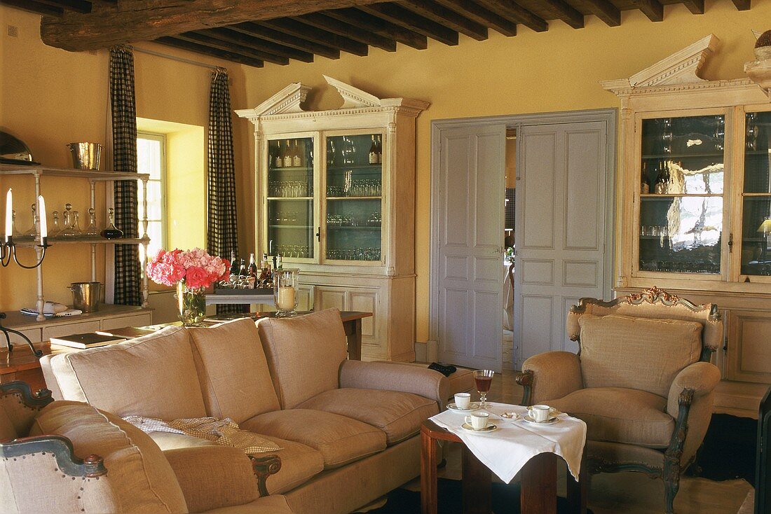 Wohnzimmer mit Flügeltür und zwei Vitrinenschränken, weich gepolstertem Sofa und Sesseln; davor ein runder, gedeckter Kaffeetisch mit weisser Tischdecke