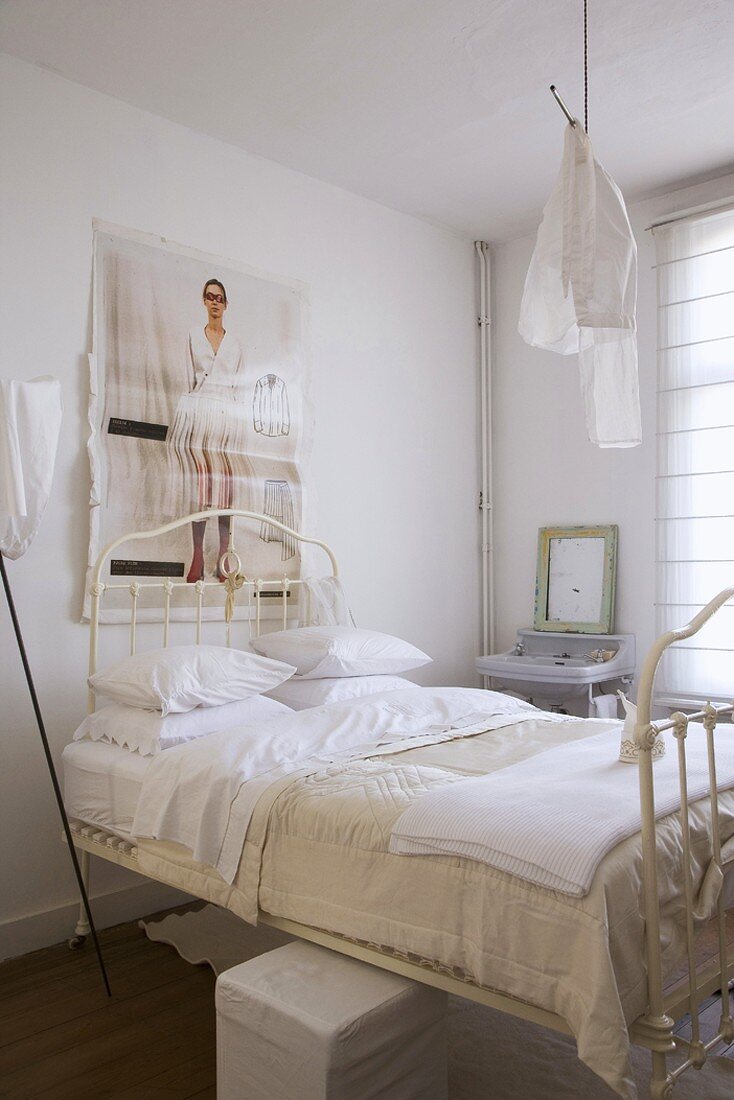 White metal bed in white bedroom with old sink, vintage mirror and cube pouffe under bed