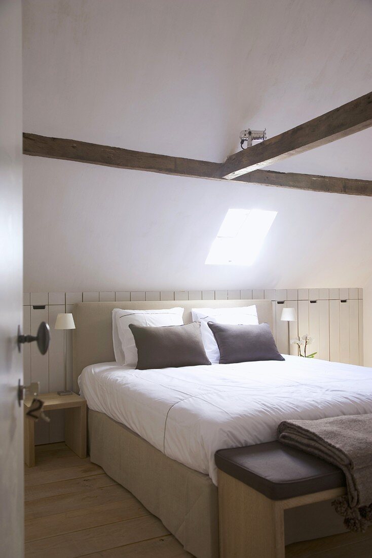 Heller Schlafraum mit Dachluke in der Dachschräge und Deckenbalken; darunter Doppelbett mit gepolstertem Kopfende