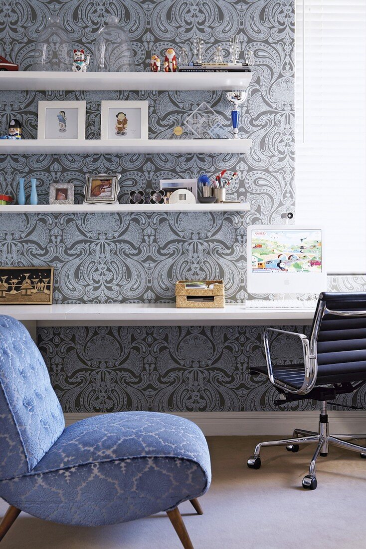 Blue patterned easy chair in front of workspace with patterned wallpaper, floating shelves and desk