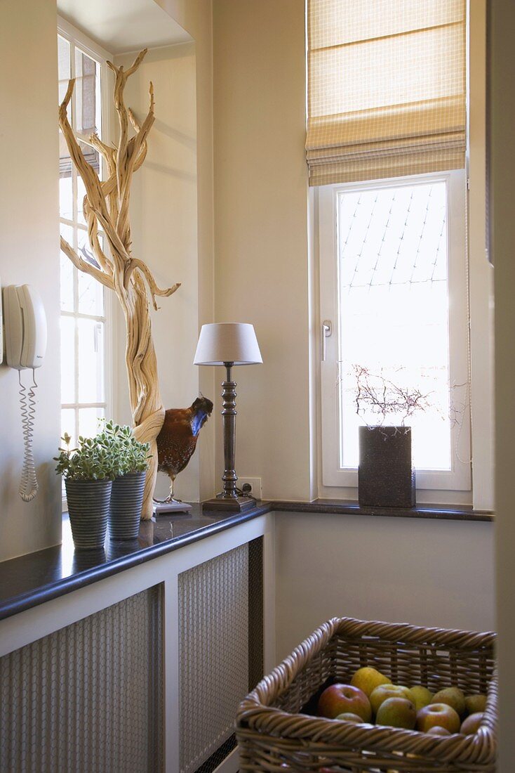 Corner of room with two windows, potted plants, driftwood and stuffed bird on windowsill and wicker basket of fresh fruit