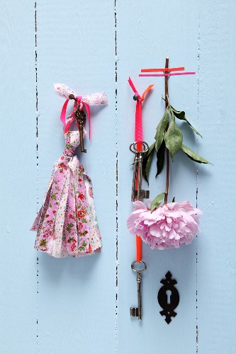 Old keys with ribbons and hand-crafted tassels made from fabric remnants hanging on pale blue wooden wall; peony stuck to wall with washi tape