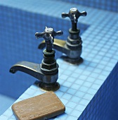 Two antique taps and bar of soap on edge of a blue tiled bathtub