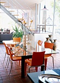 Orangefarbene Kunststoffstühle an einem Glastisch in heller zeitgenössischer Architektur mit offener Treppe
