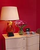 Table lamp with glass base and vase of pink anemones against magenta wall