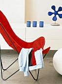 Schmetterlingsstuhl mit Sportjacke über der roten Sitzfläche und weisses Sideboard mit blauer Deko