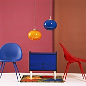 Kleine blaue Kommode zwischen Kunststoff-Schalenstühlen und zwei Retro-Hängelampen aus farbigem Glas