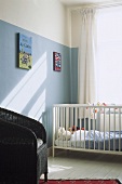 Cot below window and wicker chair against wall painted pale blue