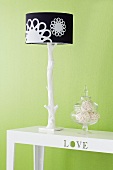Table lamp with black and white lampshade and natural wood-style base next to glass jar on table against green-painted wall
