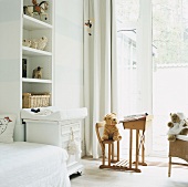 Freundliches Kinderzimmer in Weiß mit Stofftieren, Spielzeug, einer Wickelkommode und einem raumhohen Fenster