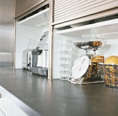 Arbeitsfläche einer modernen Küche mit neuwertigen Küchenutensilien und -geräten, gestapelten Gläsern und einer Obstschale aus Edelstahl