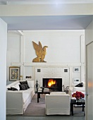 Weisses Wohnzimmer mit weisser Sitzgarnitur, brennendem Kaminfeuer und einer goldenen Adlerfigur auf dem Kaminsims