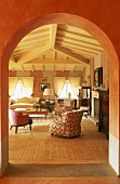 Blick durch einen Rundbogen auf terrakottafarbenen Wohnraum mit Balkendecke, offenem Kamin und gemütlichen Polstermöbeln