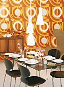 Gedeckter Esstisch und Kommode mit Käsefondue-Set vor Wandtapete im 70er Jahre Stil