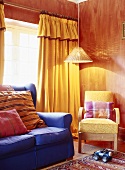 Blaues Sofa und gelber Sessel unter leuchtende Hängelampe im Wohnzimmer mit roten Wänden und gelben Vorhänge