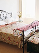 Romantisches Doppelbett aus Metall und eine florale Bettdecke, daneben ein kleiner Nachttisch mit Retroleuchte