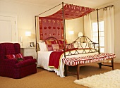 Ein Himmelbett aus Metall und ein schwerer Polstersessel im rot akzentuierten Schlafzimmer